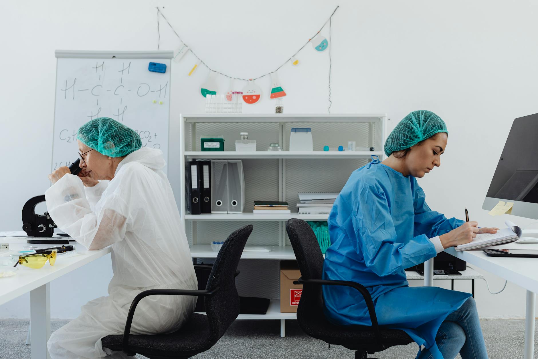 Two scientists in a lab, one examining a microscope, the other taking notes. (Photo by www.kaboompics.com on Pexels)